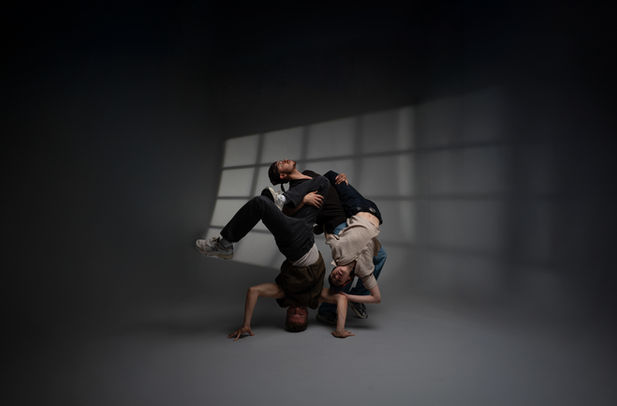 Drei Personen machen in einem leeren Raum vor einer grauen Wand eine Pose, die an Breakdance erinnert.