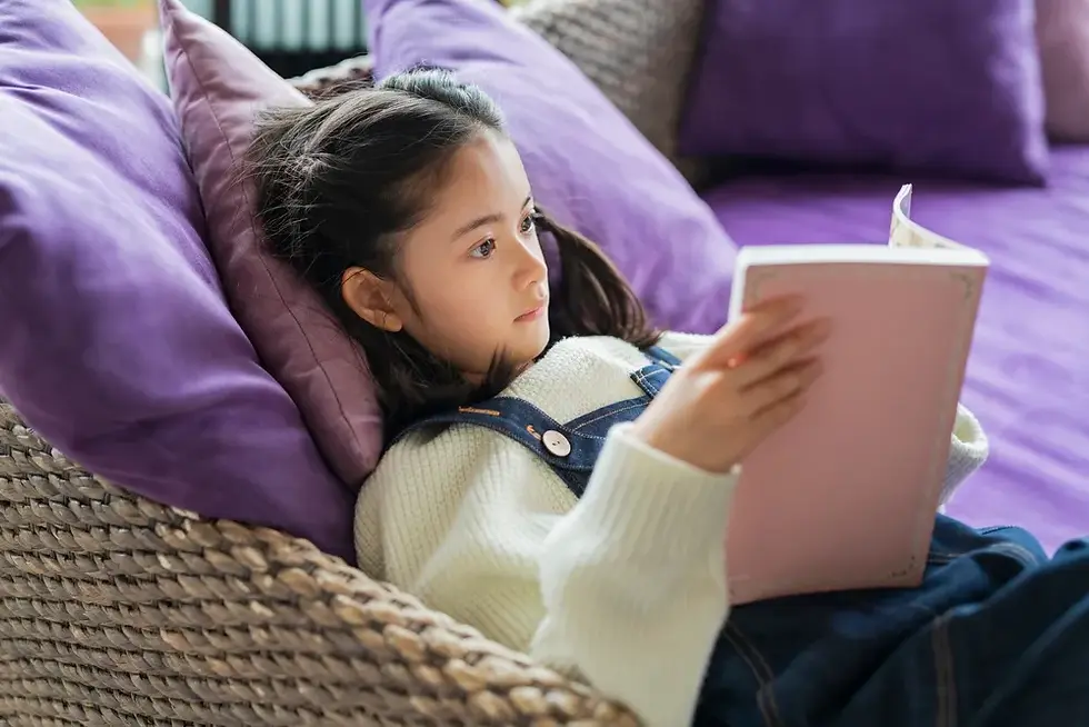 A young girl comfortably reclines on a cushioned chair, deeply engrossed in her book, as the soft light casts a cozy ambiance around her.