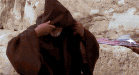 A cloaked figure lifts a hood against a rocky desert backdrop. The atmosphere is mysterious, with muted earth tones dominating the scene.