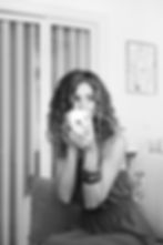 Woman with curly hair smiling, holding a "Be Happy" mug. Black and white photo in a cozy room with a modern art piece on the wall.