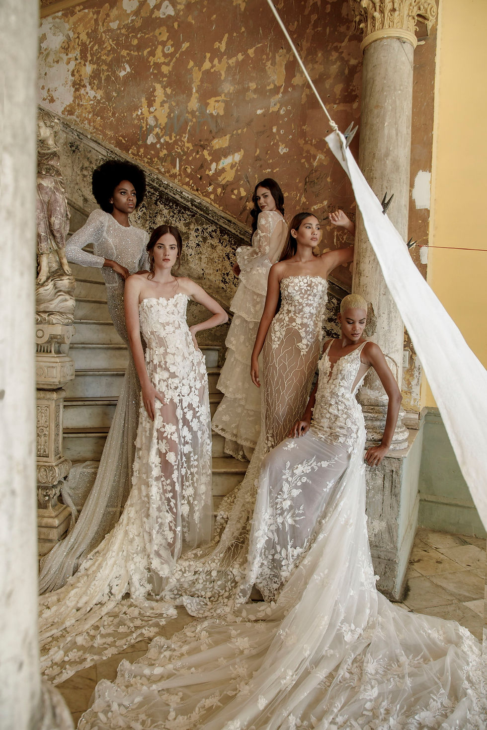 Five brides posing on an old staircase of a castle, all with laced and semi transparent wedding gowns