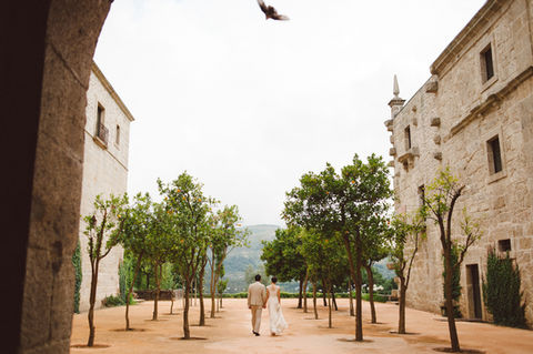 Bride and groom walking hand in hand between orange trees in the patio of an old and fully recovered monastery