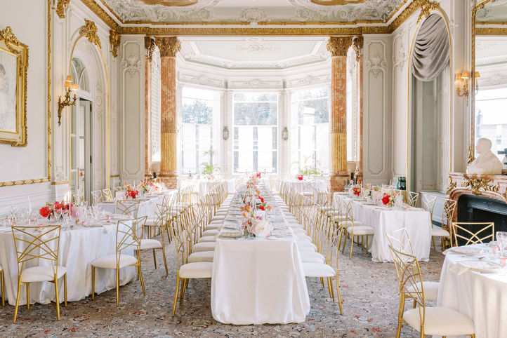 Majestic dining room with golden Pilars and baroque ceiling. Round and rectangular tables with white tablecloths and golden chairs. Small colourful floral arrangements on each table