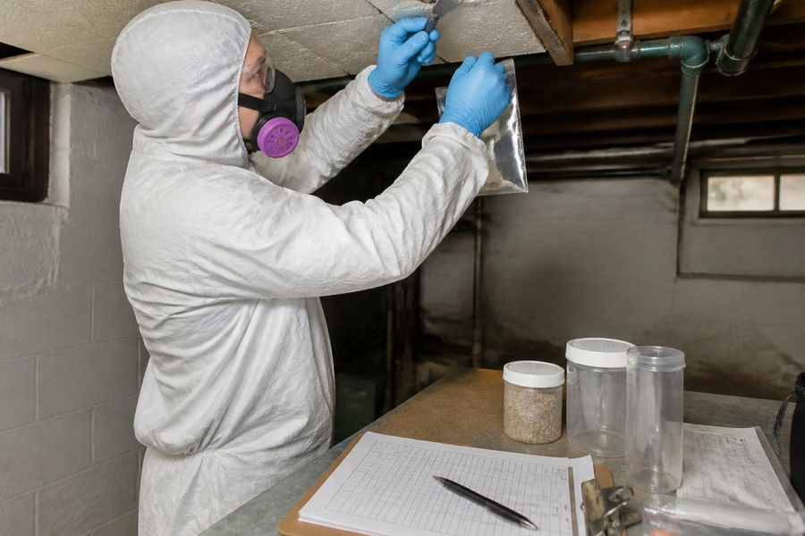 Professional asbestos inspector in white protective suit and gloves carefully collecting a bulk sample from suspect ceiling tile material in a basement, with sample containers, chain-of-custody forms, and documentation clipboard visible on a nearby work table