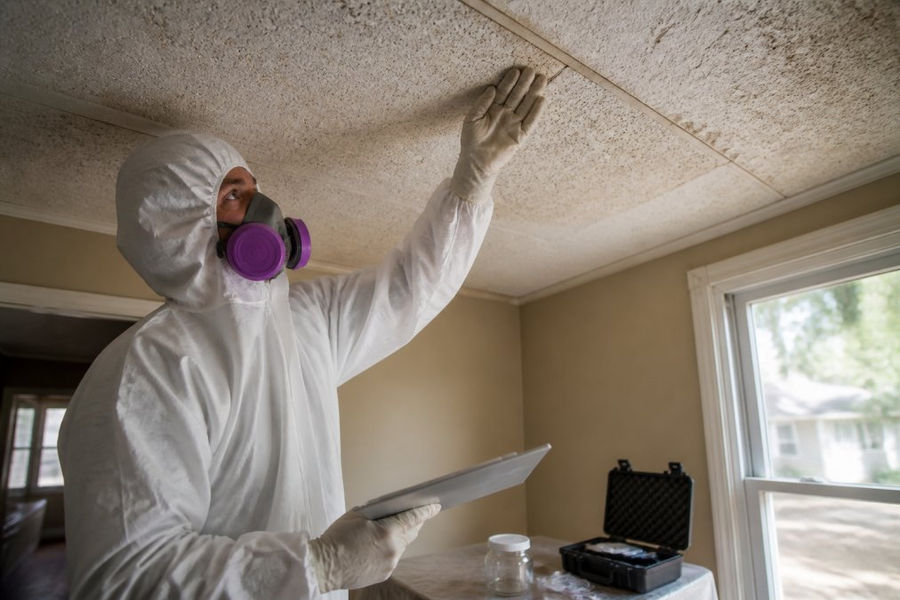 Certified inspector in protective gear examining a ceiling tile for a second asbestos inspection in an old home