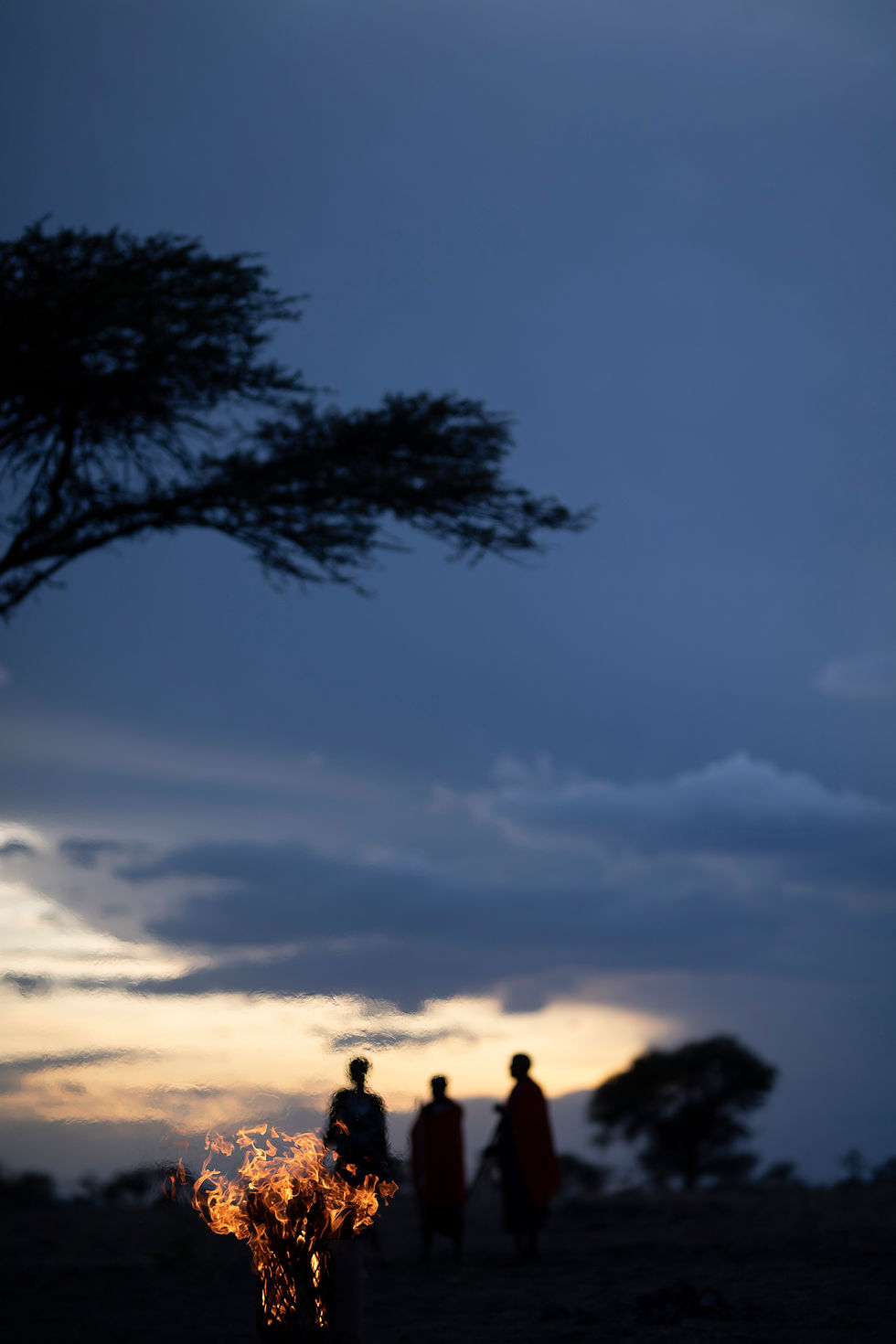 Maasai silhouettes standing near fire at sunset in Tanzania, documentary adventure photography by Michal Dzikowski