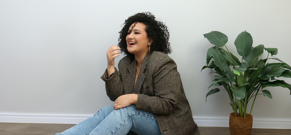 A woman with curly hair, wearing a patterned blazer and jeans, sits on the floor next to a potted plant, laughing with her mo