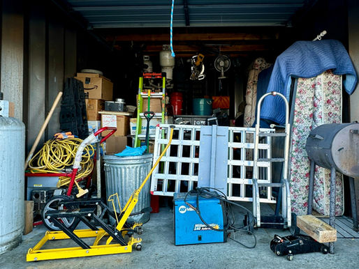 Cluttered garage with tools, propane tank, cables, ladders, and boxes. A blue tarp covers floral fabric; welding machine visible.