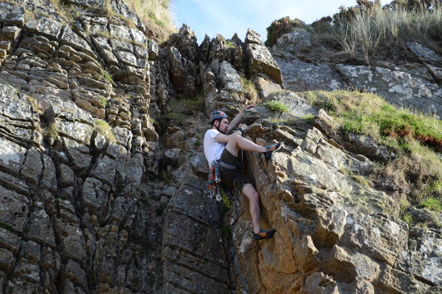 James Iain Sword climbing on a cliff performing the final move of a trad climb