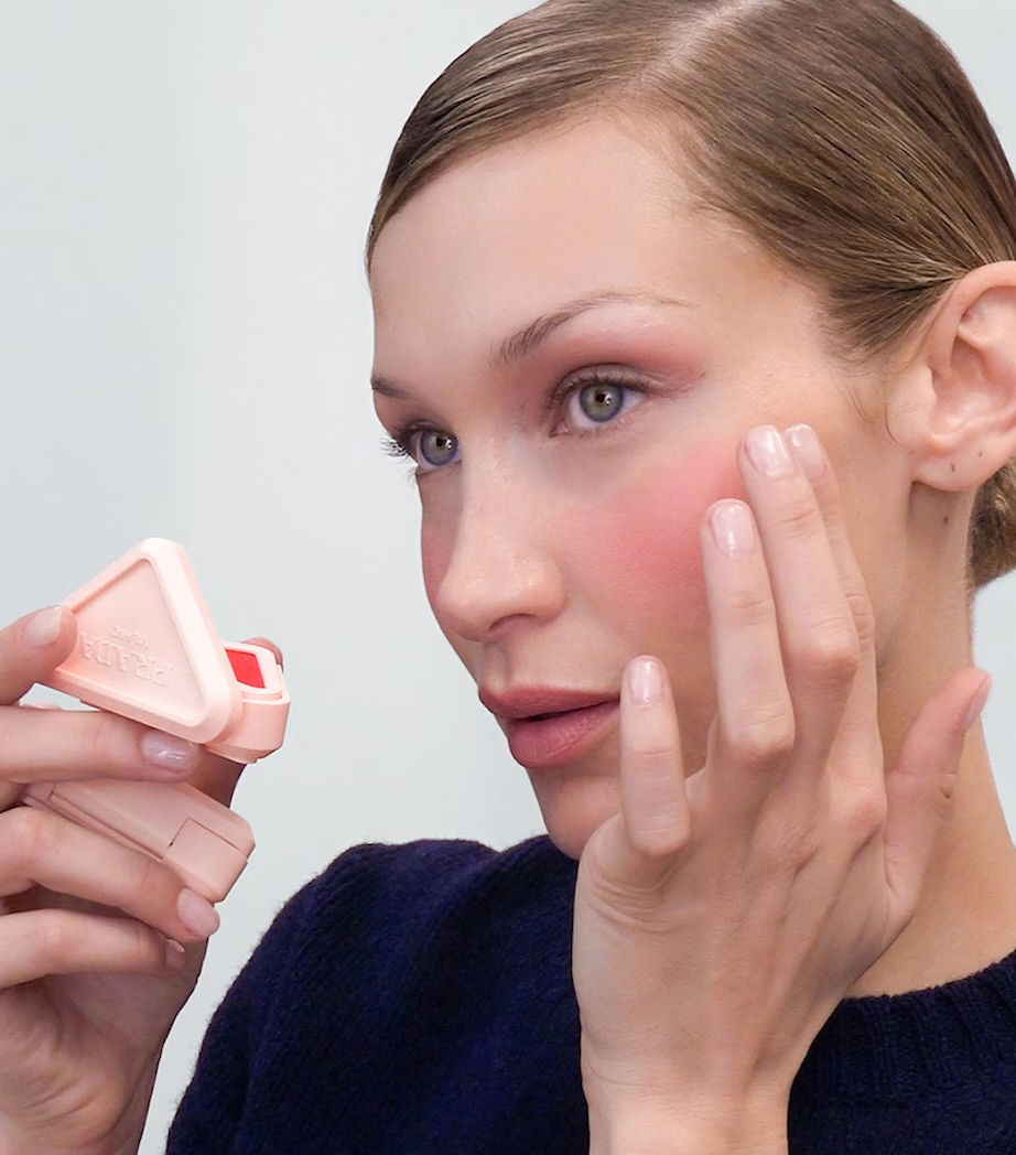 Woman applying blush with fingers, holding a pink compact in a minimal setting. Soft, natural makeup look. bella hadid