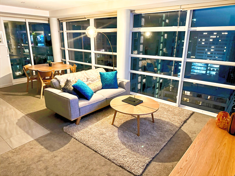 Pinnacle Apartments feature modern living room with large windows showing a city skyline at night. Gray sofa with blue pillows, wooden table, and warm lighting. Cozy ambiance.