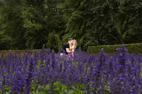 A couple shares a tender embrace amid a field of vibrant purple flowers at Nemours Estate. The woman, dressed in a flowing pink dress, gently holds her partner’s face as they smile, surrounded by lush greenery. The blurred foreground of blossoms adds depth, enhancing the romantic and dreamy atmosphere.