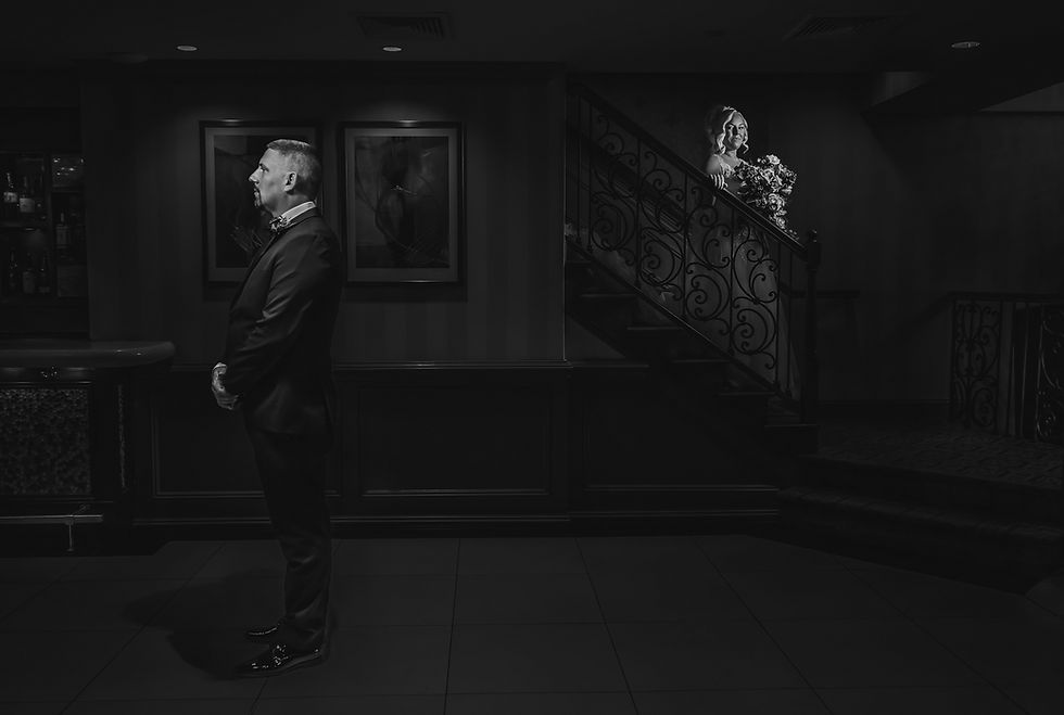 A bride walking down the stairs with a smirk on her face. A groom standing and waiting for his bride to arrive. A dramatic black and white setting.