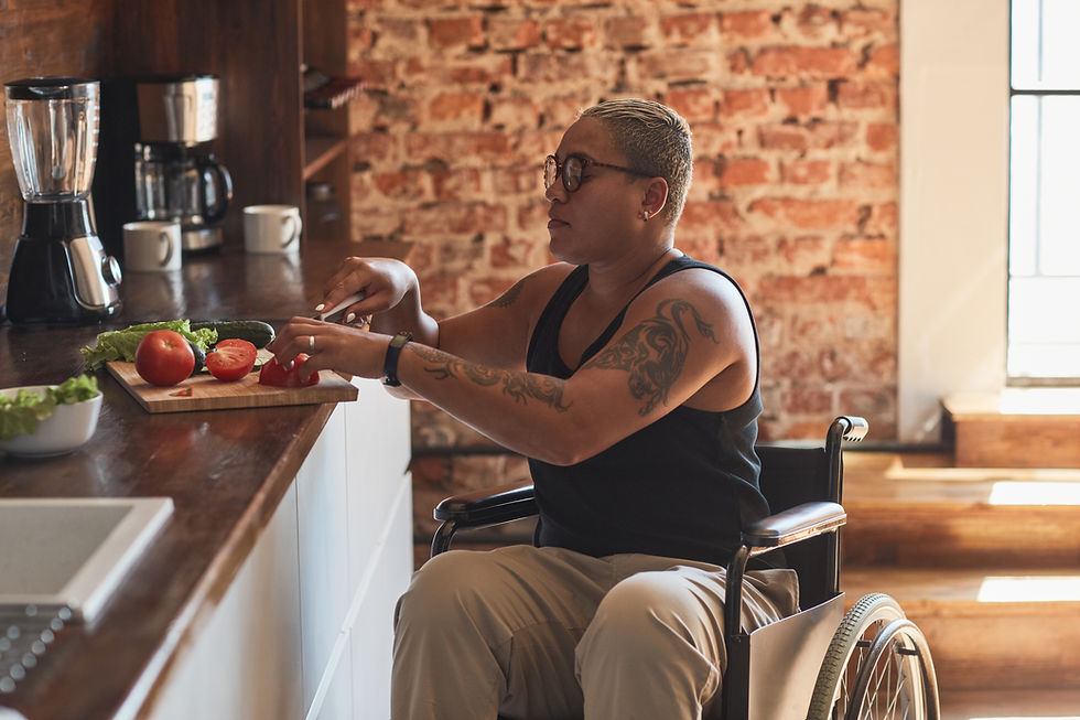 Woman in a wheelchair chops tomatoes in a warm brick-walled kitchen with lettuce, cucumbers, and a blender on the counter.