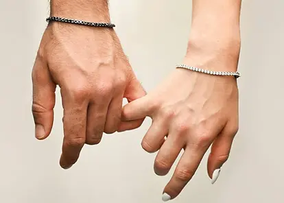 Dos manos entrelazadas, una de hombre y otra de mujer, luciendo pulseras riviere finas: una con piedras oscuras y otra con piedras claras, sobre fondo neutro