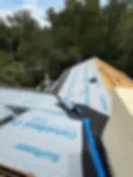 Roof with blue CertainTeed underlayment, surrounded by trees. Rolled materials and tools on surface, blue sky visible above.