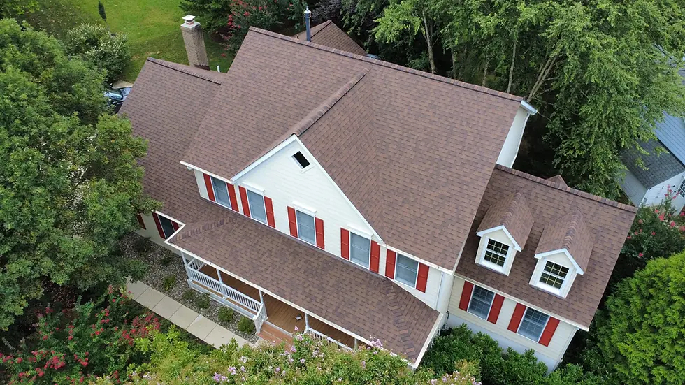 Aerial view of a house with brown roof, red shutters, and white siding. Surrounded by lush green trees and a pathway, creating a peaceful setting.