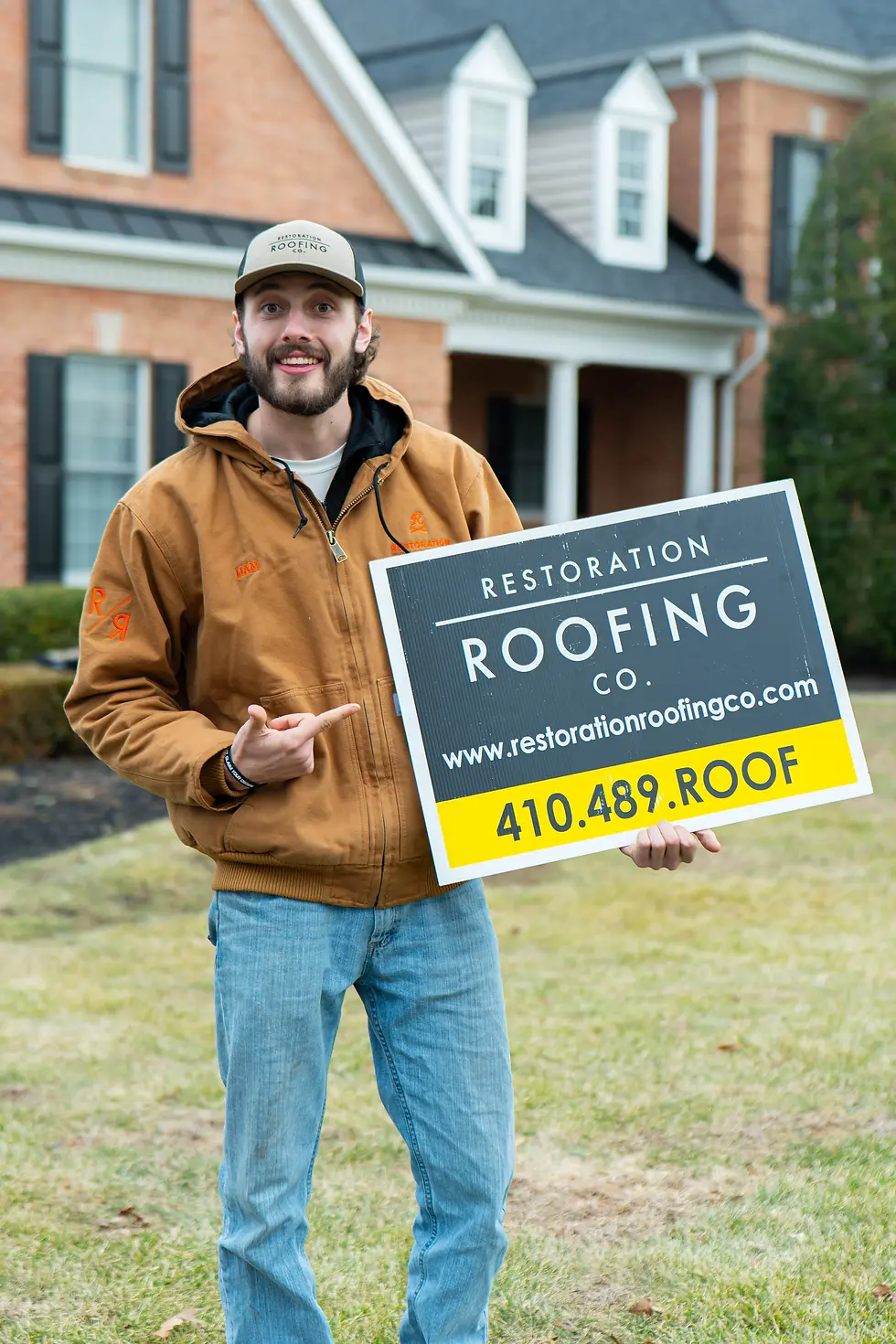 Man in a brown jacket holds a "Restoration Roofing Co." sign with contact details. He's smiling in front of a brick house.