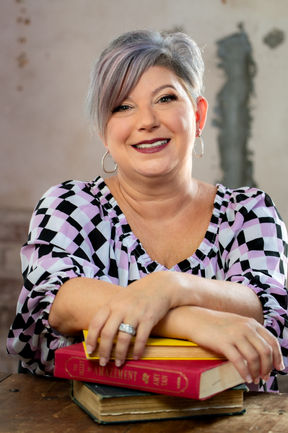 Woman with short gray and purple hair smiles at the camera with her arms crossed over a stack of books