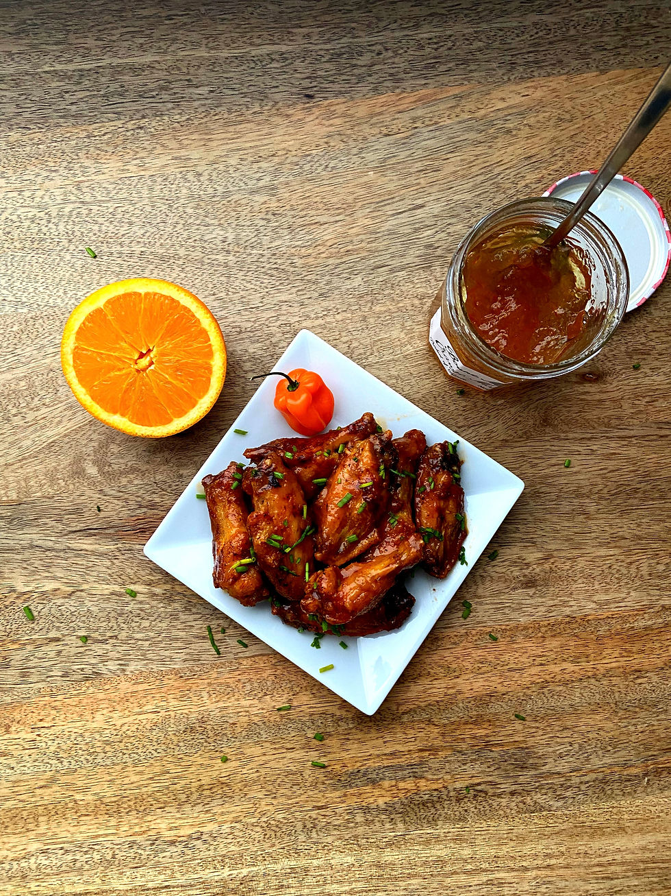 Wings on a white plate with a habanero next to it all on a wood table next to half an orange and an open jar of orange marmalade