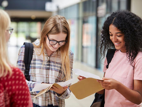 Two teenage girls opening their GCSE results in front of a teacher at schoo.