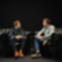 two younger men talking sat on Chesterfield sofas dark plain background.jpg