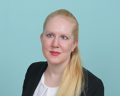 Against a blue background, a very fair woman with long blonder hair smiles. She wears a black blazer and white blouse.