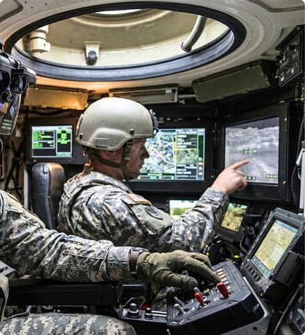 Inside the cramped, metallic interior of an armored military vehicle, a soldier wearing a combat helmet and camouflage uniform points to a digital touchscreen monitor displaying a thermal imaging feed. In the foreground, another soldier's gloved hand rests on a control panel.