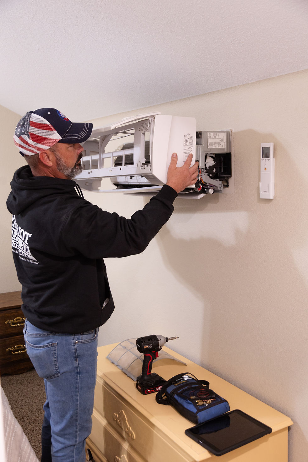 Patriot Heat & Air technician installing a ductless mini-split unit inside a home, wearing a branded hoodie and American flag hat.