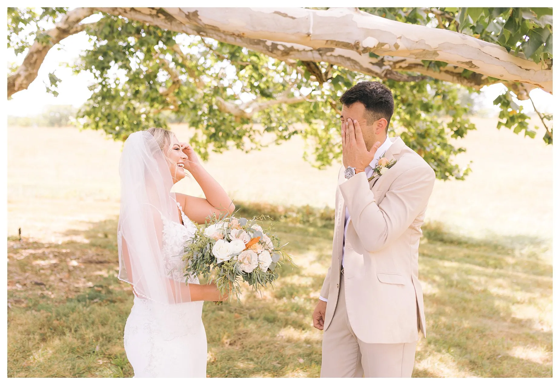 Bride and groom under a tree, both smiling and covering eyes. Bride holds bouquet. Sunlit outdoor setting with green foliage.