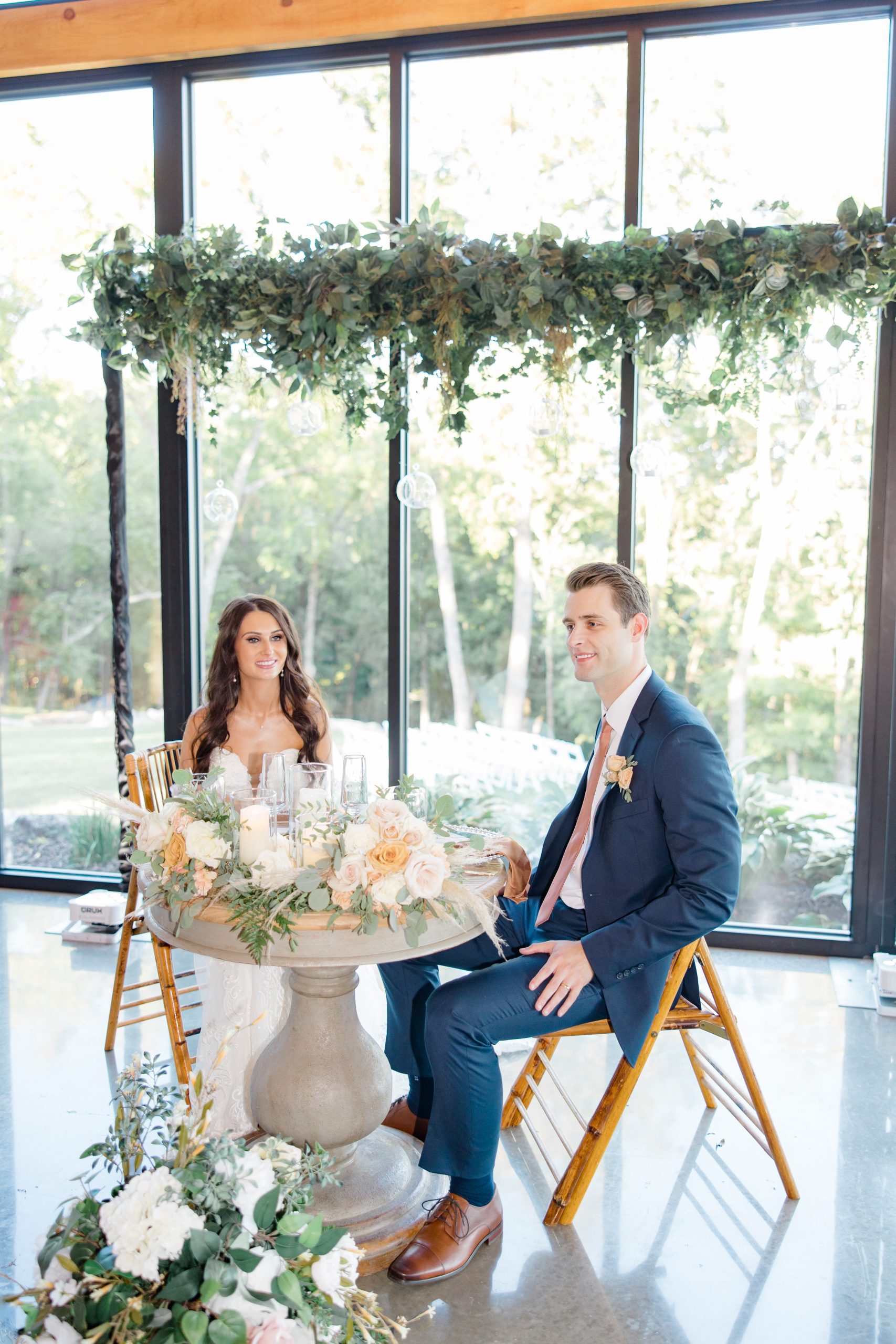 A couple sits at a round table decorated with pastel flowers. Large windows show a green garden. The mood is joyful and serene.