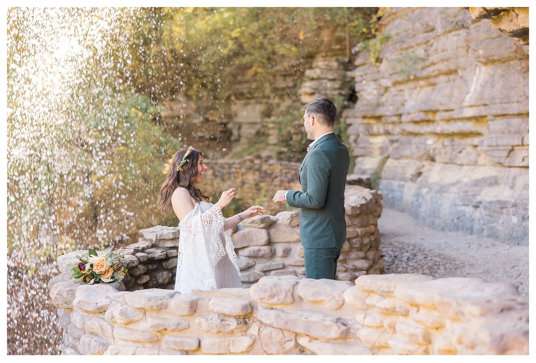 First Look with a Bride and Groom underneath a Waterfall at Dogwood Canyon. The Groom is in a Green Tuxedo and the Bride is wearing a white lace boho wedding dress and a floral crown.