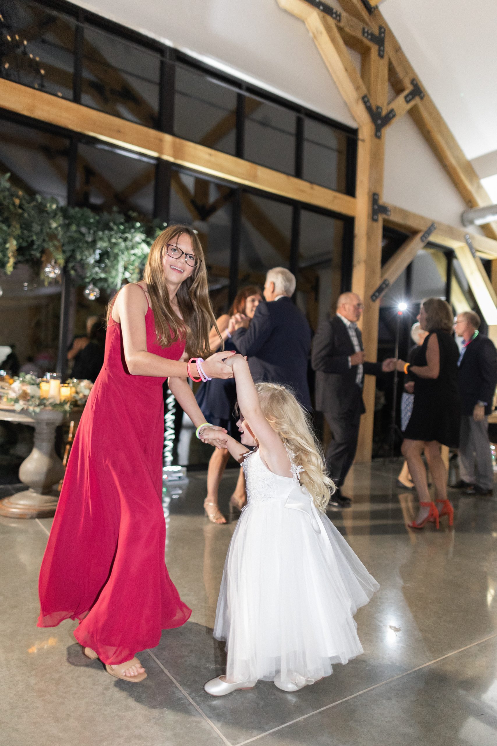 Woman in a red dress and a girl in white dance joyfully at an indoor event with a wooden beam ceiling. Others dance in the background.