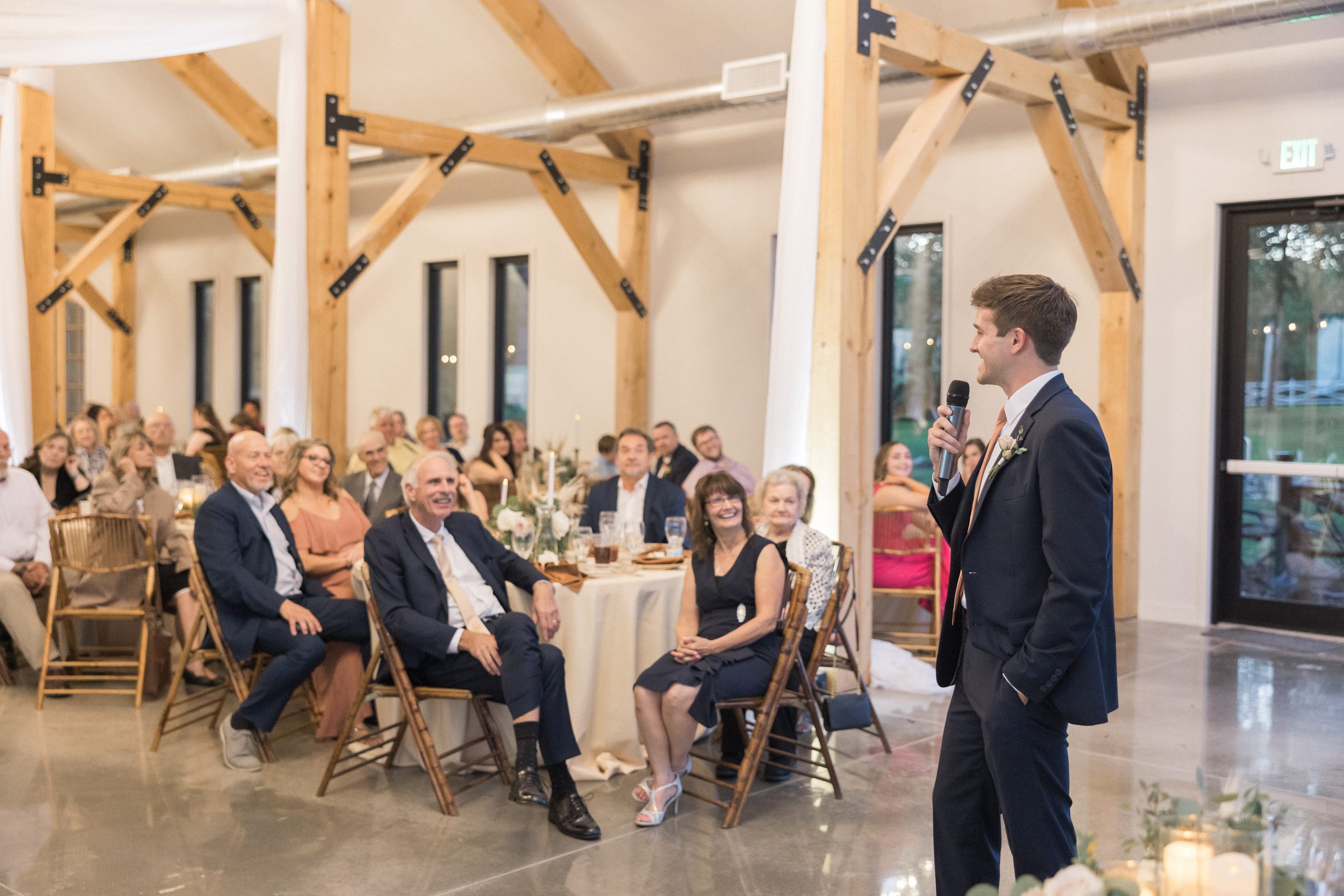 Man in a suit speaks into a mic at a wooden-beamed venue. Attentive audience sits at tables with flowers and candles, creating a joyful atmosphere.