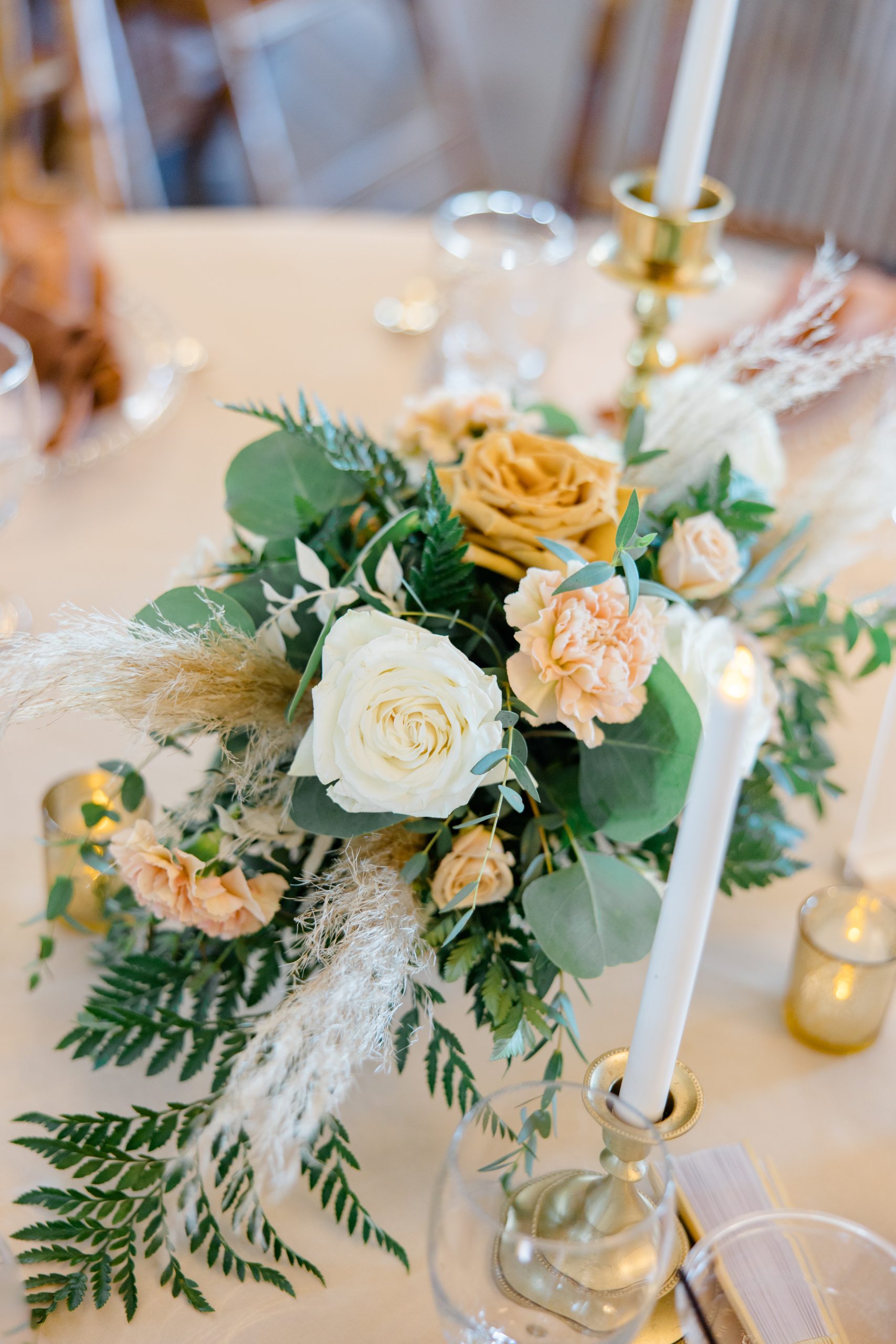 Elegant floral centerpiece with white and peach roses, greenery, and candlesticks on a table. Warm, inviting setting with a cozy feel.