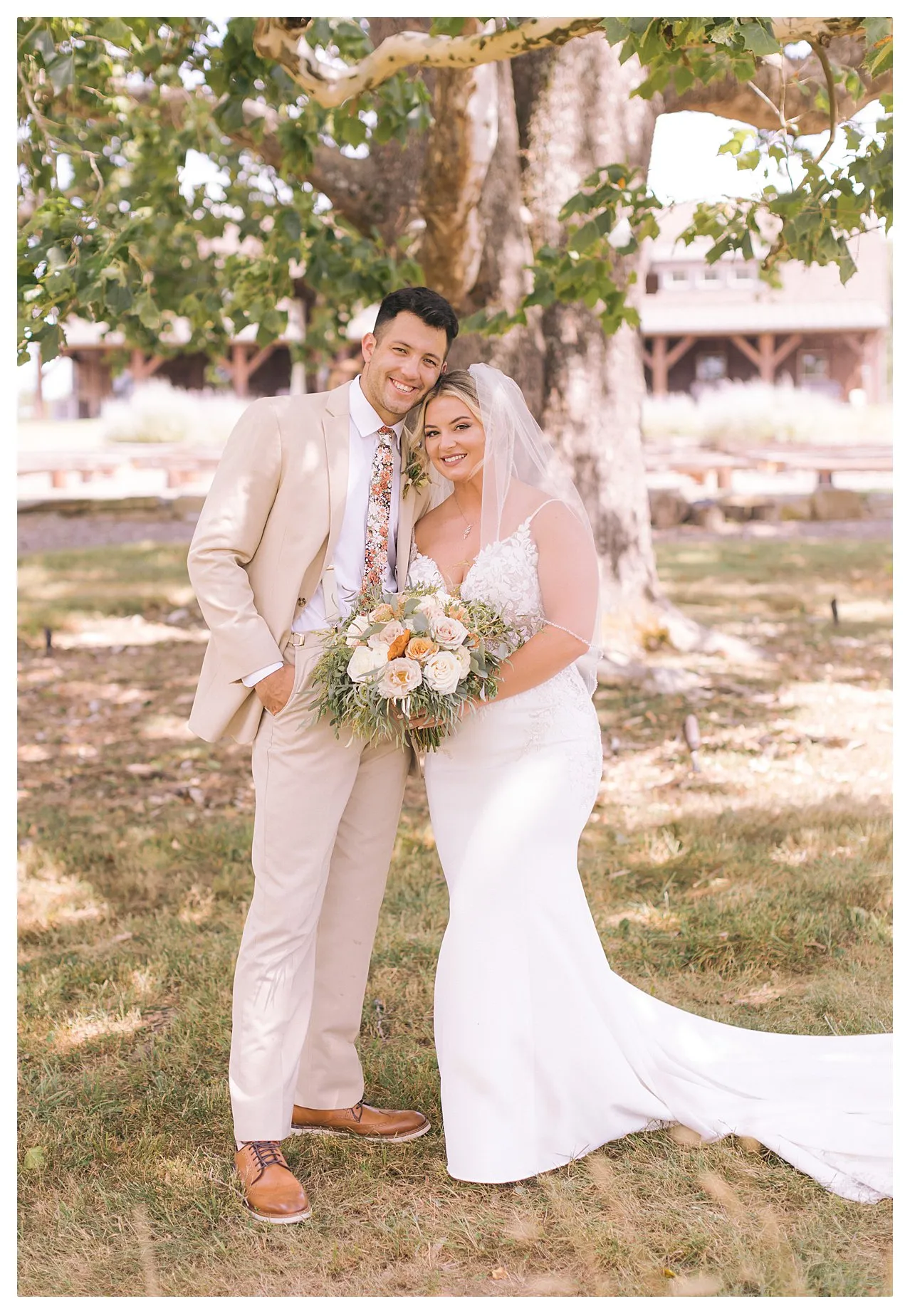 Bride and groom smiling under a tree. Bride holds a bouquet, wearing a veil and white dress. Groom in beige suit. Outdoor setting.