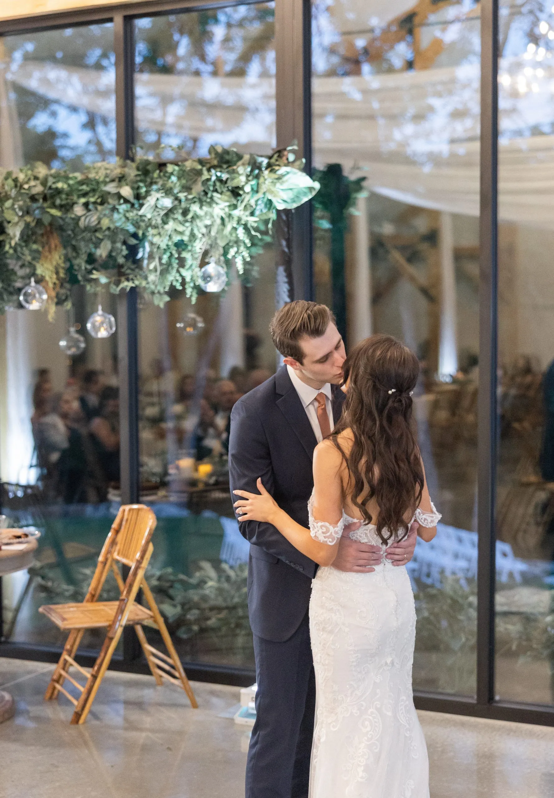 Bride and groom share a kiss under greenery in an elegant venue. She wears a lace dress, he a suit with a copper tie. Romantic ambiance.