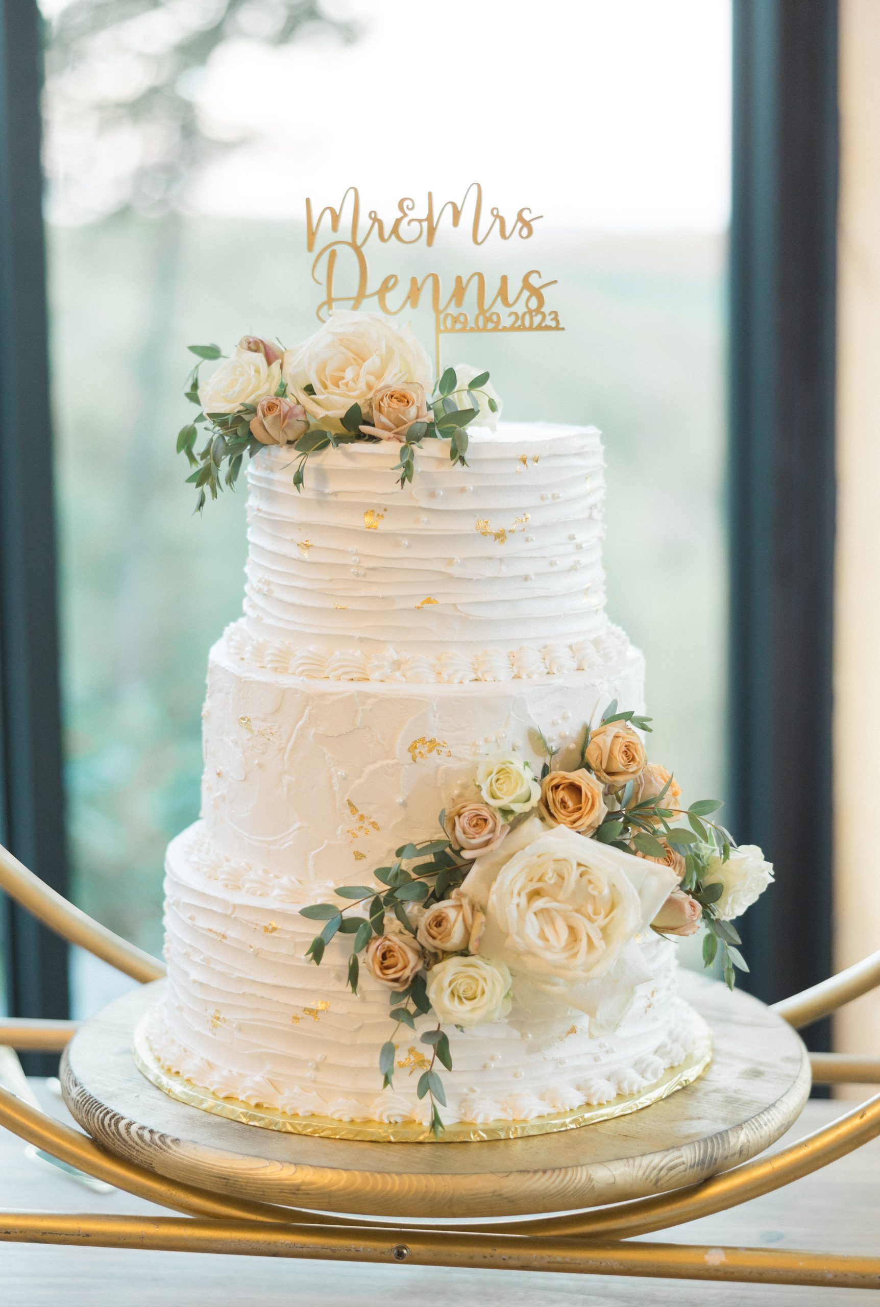 A white tiered wedding cake with gold accents, topped with roses and greenery. "Mr & Mrs Dennis 09.09.2023" displayed on top, set by a window.