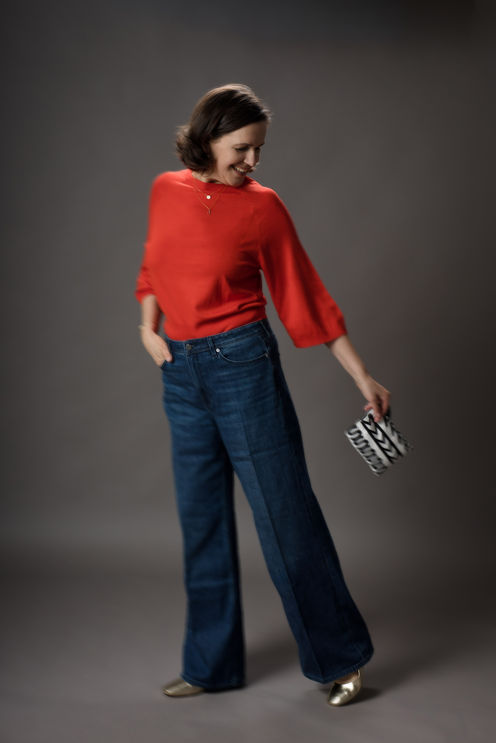 Frau in rotem Pullover, blauen Schlagjeans, hält gemusterte Clutch, lächelt.
