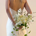 Bride in a minimalist, backless white gown holding a delicate bouquet of white and pale yellow flowers with airy greenery, styled in a natural, organic shape.