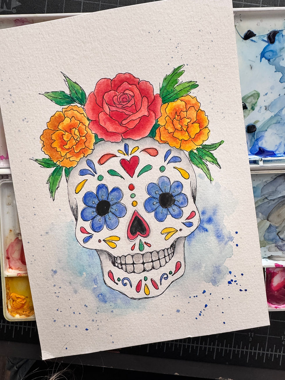 Watercolor painting of a sugar skull with a red rose, orange marigolds and blue flowers, green leaves, with a splatter background.