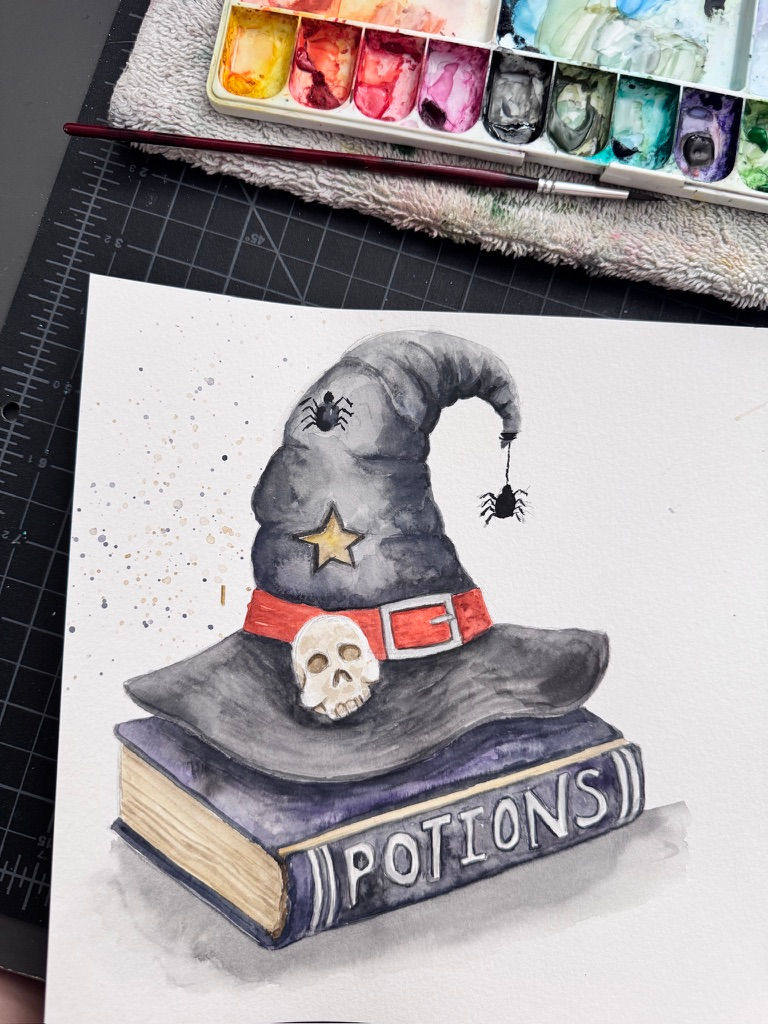 Watercolor of a spooky black wizard hat with skull and spiders on a book labeled POTIONS, beside a paint palette.