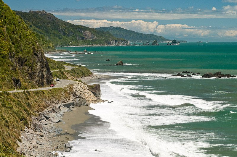 Cliffside road along a rugged coast with turquoise waves, rocky beach, and distant mountains under a bright blue sky.