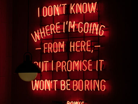 Red neon sign reads "I don't know where I'm going from here but I promise it won't be boring" against a dark background with a lamp.