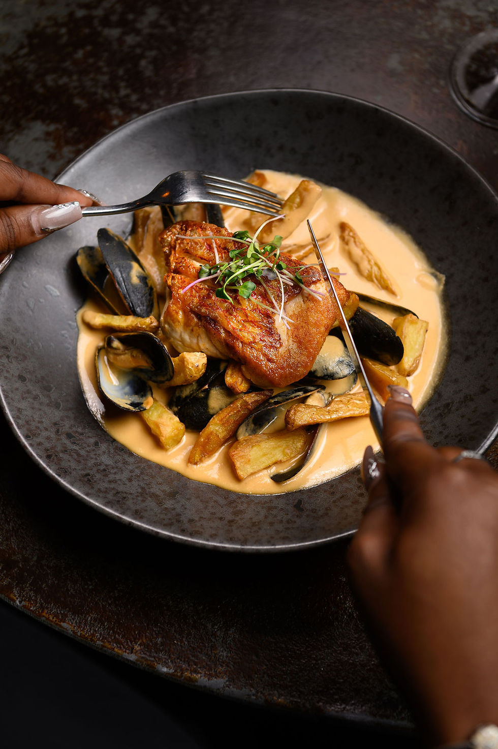 an elegant black bowl with seared snapper fillet, mussels, and a rich creamy apple sauce, a bottle of wine and a glass also on the table