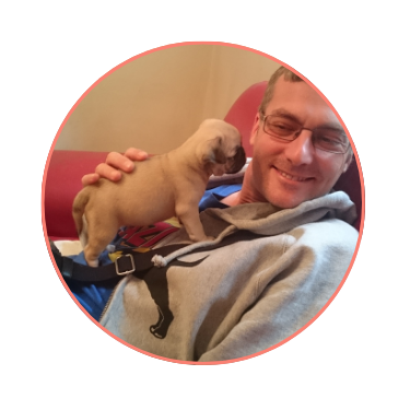 Man smiling while cuddling a small puppy on his lap, sitting on a red couch. The scene conveys warmth and happiness.