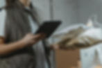 Person in a vest holds packages and a tablet in a dimly lit room with a computer in the background. Focused, efficient atmosphere.