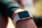 Close-up of a smartwatch on a person's wrist showing 6:13 with notifications. Rose gold with a white strap. Blurry background.