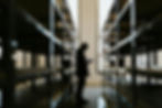 Silhouette of a person writing on a clipboard stands between metal shelves in a dimly lit warehouse. Light streams through a window.