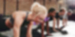 Four people in a gym doing push-ups on a black mat. They're focused and determined, with a brick wall in the background.